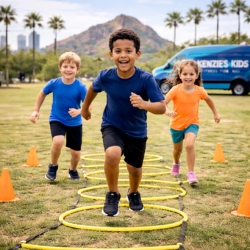 Three smiling children run agility ladder drills on a grassy field during a Kenzie's Kids mobile fitness session in Phoenix, AZ. With palm trees, city skyline, and a branded Kenzie's Kids van in the background, the scene promotes active play, coordination, and confidence.