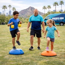 A male Kenzie's Kids trainer guides a young boy and girl during a functional fitness session in Phoenix, AZ. The children perform balance drills on stability pods while holding kettlebells, with a city skyline, palm trees, and a branded mobile gym van in the background.