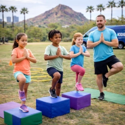 A male Kenzie's Kids trainer leads three diverse children in a balance and mindfulness exercise on foam blocks during a mobile fitness session in Phoenix, AZ. Set against a backdrop of palm trees, desert mountains, and a branded van, the activity promotes emotional regulation and focus through movement.