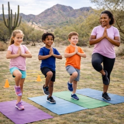 A diverse group of three children and a female fitness coach practice yoga Tree Pose on colorful mats in a Tucson, AZ desert setting. The scene features mountains, cacti, and cones in the background, promoting movement for emotional regulation and focus.