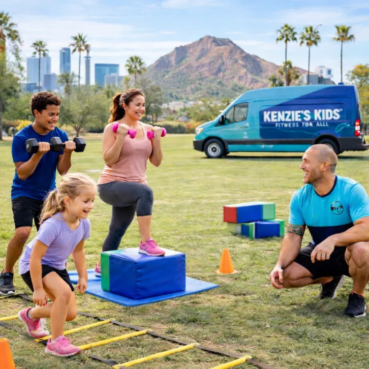 Children and adults participate in an outdoor mobile fitness session in Phoenix, AZ. A woman performs step-ups with pink dumbbells on a foam block while a young girl does agility ladder drills and a teen lifts dumbbells. A Kenzie's Kids coach supervises near a branded van, with palm trees, city skyline, and mountains in the background.