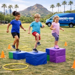 Three children step across foam blocks during a Kenzie's Kids functional fitness session in Phoenix, AZ. The activity promotes coordination and progressions, set on a grassy field with agility rings, cones, palm trees, and a branded mobile gym van in front of a desert mountain backdrop.