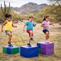 Three children perform step-up exercises on colorful foam blocks in a desert park in Tucson, AZ. The diverse group of kids practices strength and coordination with agility rings and cones in the background, set against a scenic mountain and cactus landscape.