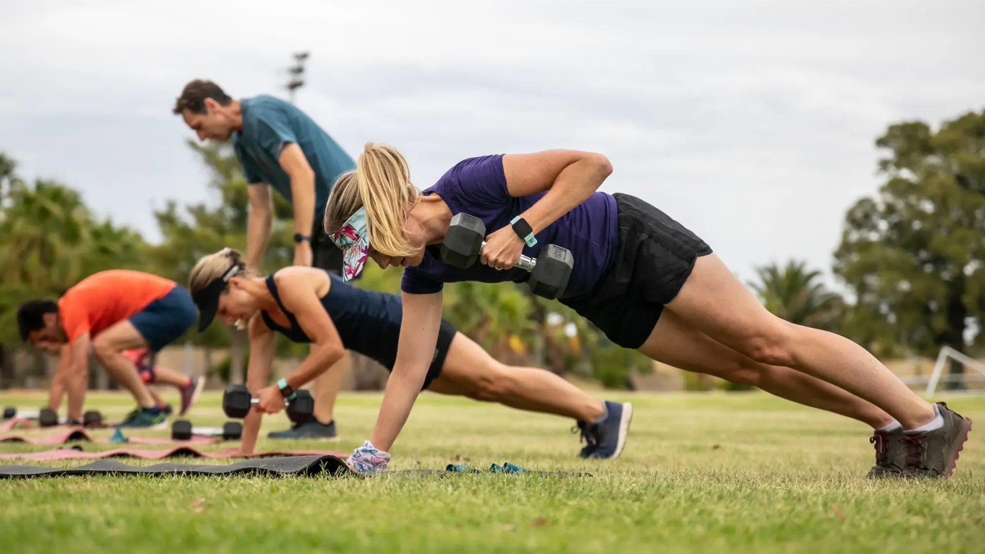 Adults performing outdoor strength exercises with dumbbells during a group fitness session