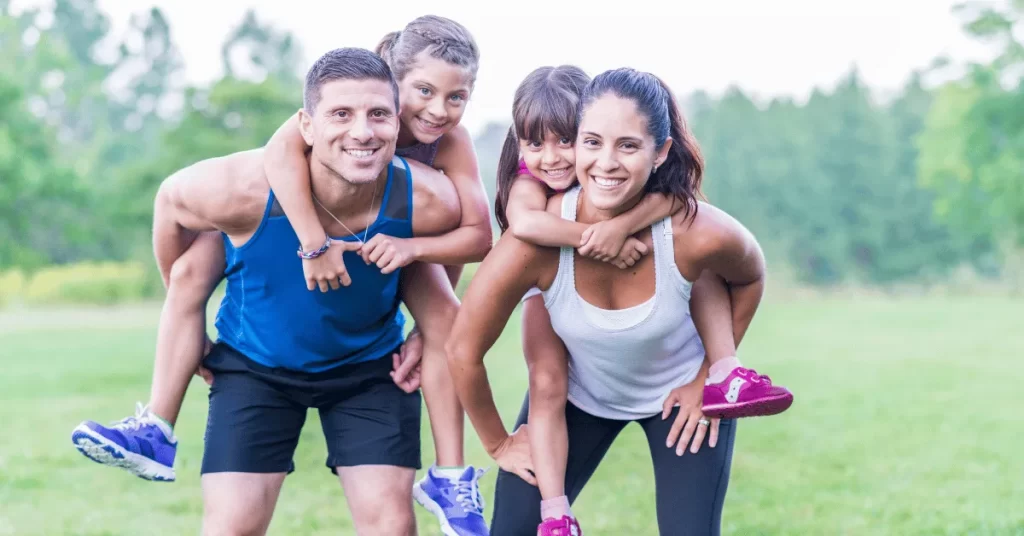 Family exercising outdoors with parents carrying kids on their backs, smiling and staying active together in a park setting
